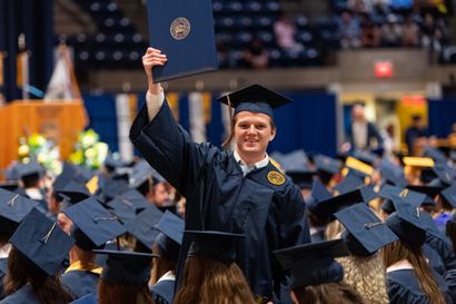 A male graduate holds up his diploma amid a crowd of other students, all wearing their cap and gowns