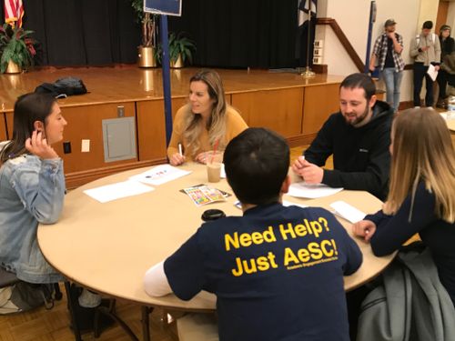 "Need help just aesc" quote on back of t-shirt, group of students sitting at table